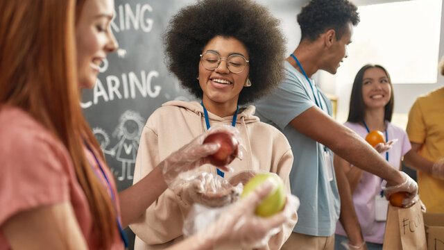 Happy young volunteers in gloves collecting, sorting food for needy people, Diverse team working together on donation project in charitable organization office