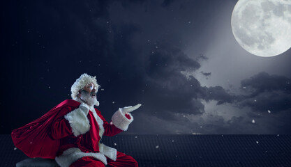 Fototapeta premium Emotional Santa Claus congratulating with New Year 2021 and Christmas. Man in traditional costume sitting on house roof with full moon on background in midnight. Winter, holidays, sales. Copyspace.