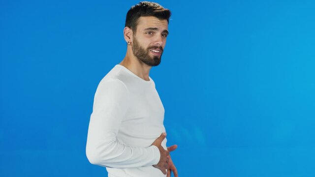 Male suffering from digestive problems. Bearded man wearing white t-shirt sudden stomach ache, severe abdominal pain, constipation, gastritis, pancreatic diseases. Blue background.