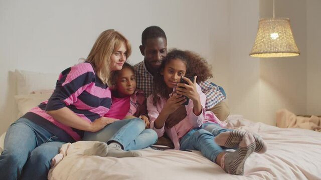 Positive smiling interracial family with two preadolescent cute mixed race daughter sitting on bed in love embrace, taking selfie shot on smart phone while enjoying leisure together in evening.
