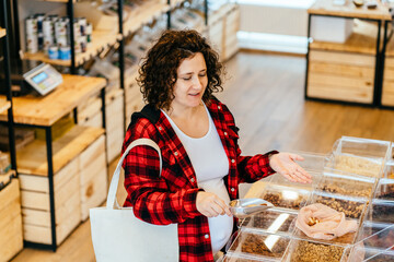 Sustainable shopping at small local businesses. Portrait of pregnant female putting nuts in cotton produce bag at plastic free store. Reusable eco bag for shopping. Zero waste concept.