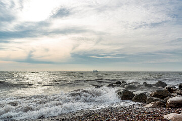 Fototapeta na wymiar Wellen treffen auf den Ostseestrand und die Wellenbrecher. Am Horizont fährt ein Schiff.