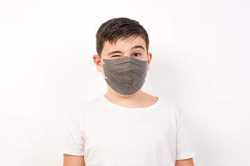 Caucasian young boy wearing medical mask standing against white background blinking eyes with pleasure having happy expression. Facial expressions and people emotions concept.