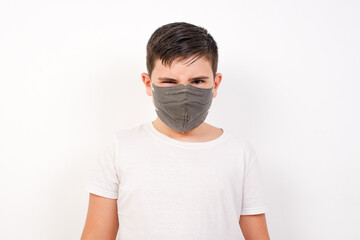 Caucasian young boy wearing medical mask standing against white background frowning his eyebrows being displeased with something.