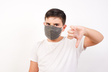 Discontent Caucasian young boy wearing medical mask standing against white background shows disapproval sign, keeps thumb down, expresses dislike, frowns face in discontent. Negative feelings.