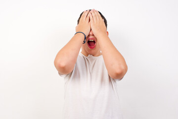 Shocked Caucasian young boy standing against white background holding hands on face covering eyes and screaming in despair and frustration while being full of terror, mouth dropped open.