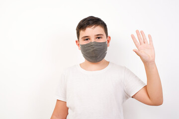 Caucasian young boy wearing medical mask standing against white background  raise one hand and wave, saying hi or hello as makes goodbye or welcome gesture.