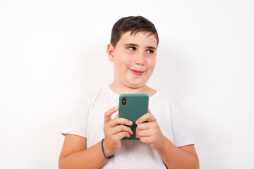 Happy Caucasian young boy standing against white background listening to music with earphones using mobile phone.