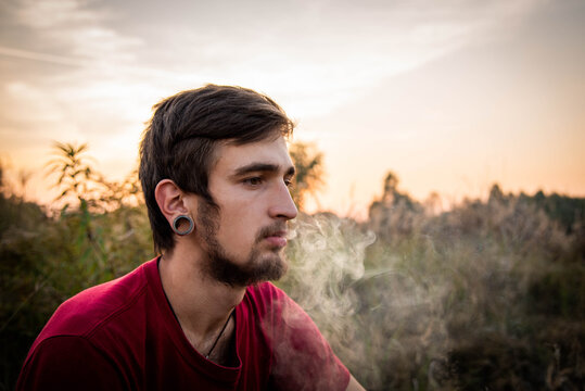 young caucasian man smoking joint with weed in the fields, growing weed plants at sunset