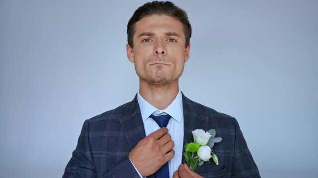 groom fixing hair, jacket and tie while looking at camera isolated on grey