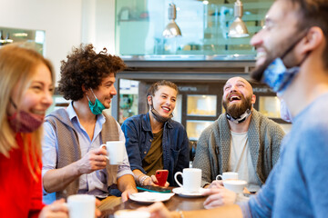 group of students having fun drinking coffee at coffeehouse wearing protective face mask. Young friends talking together at restaurant cafeteria. New normal lifestyle concept with happy guys and girls