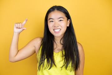 Young beautiful asian woman over yellow isolated background smiling and confident gesturing with hand doing small size sign with fingers looking and the camera. Measure concept.