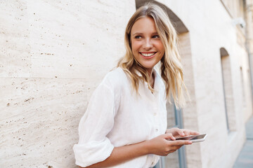 Beautiful happy woman smiling and using cellphone while leaning on wall