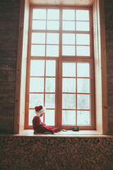 Little girl in red Santa costume is sitting by the big window with coming light playing with christmass toys