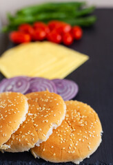 Preparation for the preparation of a hamburger, all ingredients: cheese, salad, tomato, onion, bun lie on a black stone background. Close-up.