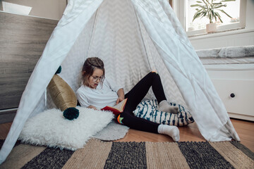 Little girl reading a book in her room in a tent.