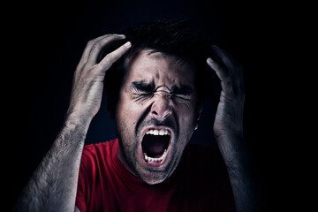 Dark short haired man in red t shirt, holding hands around his head with closed eyes screaming of pain and anger. Studio photo, dark setting.