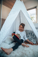 Little girl reading a book in her room in a tent.