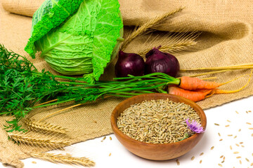 Rye seeds in wooden bowl, dry ears of rye, fresh carrot, savoy cabbage and green onion on sackcloth on white background. Organic, healthy food concept