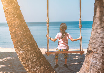 Back view of Asian child girl sitting on wooden swing at palm grove, summer vacation in Thailand,...