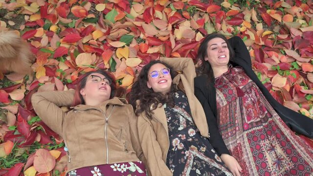 Top view three girls lying down a fallen leaves blanket with smiling faces during Autumn season. Red, yellow color, fun, friendship concept.