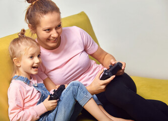 Excited young mother and cute little daughter sit on couch relax playing video games at home together, happy and small girls have fun in in living room.