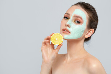 brunette woman is holding a slice of lemon in front of her face. Photo of a woman with a moisturizing face mask. Beauty and skin care concept