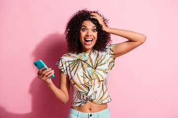 Photo portrait of excited girl holding phone in one hand touching head isolated on pastel pink colored background