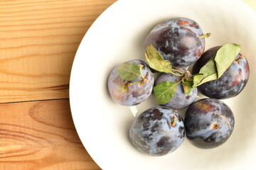 Ripe dark blue organic plums in a ceramic bowl, close-up, on a wooden table.