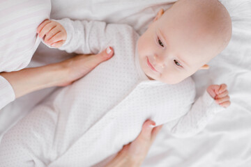 A young Caucasian mother in home clothes holds a newborn baby in a white jumpsuit. Smiles and hugs the child.