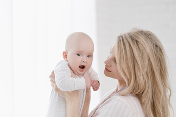 A young Caucasian mother in home clothes holds a newborn baby in a white jumpsuit. Smiles and hugs the child.