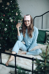 Pretty brunette girl in pyjamas and sleeping mask raising glass of champagne sitting on bed. Xmas.