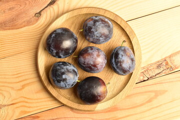 Ripe dark blue organic plums on a round wooden tray, close-up, on a wooden table.