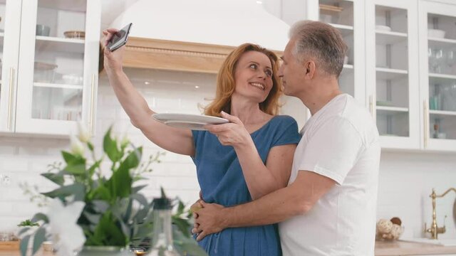 Lockdown of ginger senior Caucasian woman standing in embrace with her good-looking grey-haired husband in the kitchen and making selfie with plate of food using smartphone camera