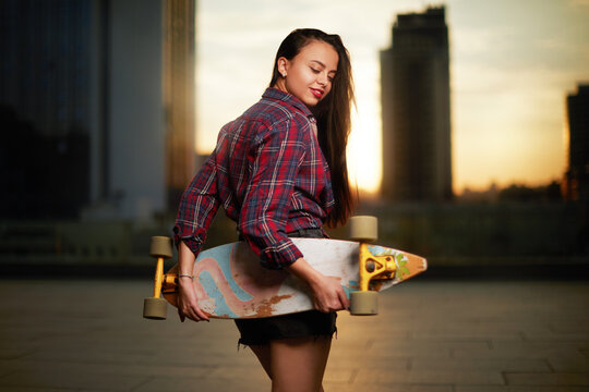 Beautiful sexy young girl in short shorts holding  longboard on sunset background in city.