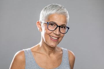 Smiling senior woman with short white hair and glasses posing in front of gray background