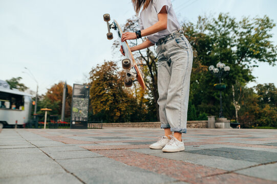 Cropped photo, woman in stylish street clothes standing on the street with a longboard in her hands. Women's skateboarding