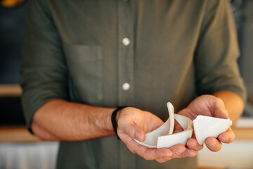 Broken ceramic white cup in the hands of a young man in the kitchen. Brokenness, failure, simplicity.