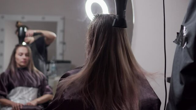 Young woman hairdresser dries with a hairdryer and combs the hair of a client in a beauty salon. Medium shot.