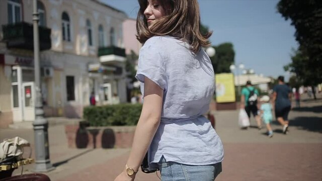 A cute young girl goes along a beautiful city square with a camera in her hand and whirling with flying hair. Medium shot. Slow motion. Blurred background