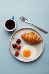 Bun, fried egg, coffee and berries at plate with fork at blue background