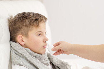 A sick child lies on the bed in a warm scarf and sprinkles medicine into his nose. Cold treatment concept