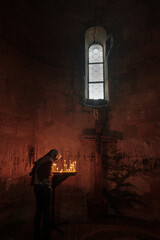 an old dark church with stone walls and a narrow small window. candles are burning in front of a cross with a rapier. a woman with a scarf on her head bowed her head in front of the cross and prays. 