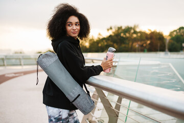 Pleased athletic sportswoman standing with water bottle and mat