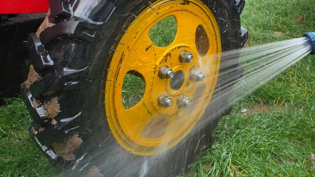 Wash truck wheels with a hose with water. Car wash. Wash the dirty tractor wheel. Water the motoblock wheel with water. Vehicle care. Vehicle washing. Running water. Human hand. Tire tread.