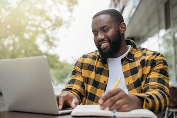 Young handsome African American man using laptop computer, taking notes, planning start up, working online. Portrait of happy student studying, learning languages, online education concept - Powered by Adobe