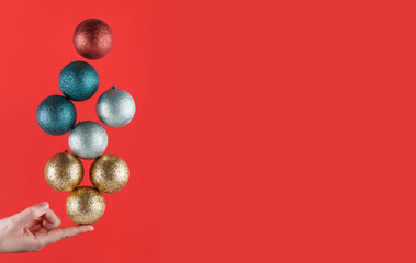 Person holding with one finger brightly colored Christmas spheres striking a balance on a red background with copy space. Christmas decorations. Christmas wallpaper
