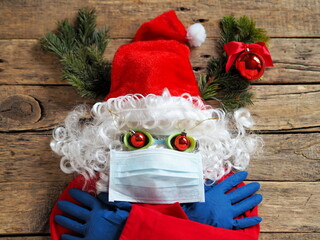 The concept of a comic image of a frightened Santa Claus in a cap made of a medical mask, gloves, glasses on a wooden background. 
