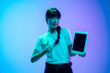 Showing tablet's screen. Young asian woman's portrait on gradient blue-purple studio background in neon light. Concept of youth, human emotions, facial expression, sales, ad. Beautiful brunette model.