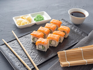 Sushi rolls philadelphia on a black textured plate stand on a gray background. wasabi ginger and sauce chopsticks. Rolls with salmon, cream cheese, avocado.